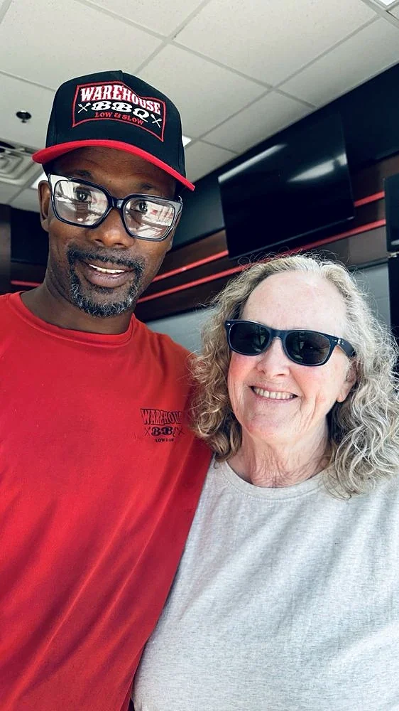 A Warehouse BBQ team member in a Warehouse BBQ cap and red shirt smiling with a guest inside the Youngsville restaurant